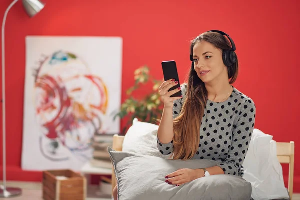 Joven morena caucásica sentada en la cama con auriculares en las orejas y usando un teléfono inteligente. Fondo rojo coral . — Foto de Stock