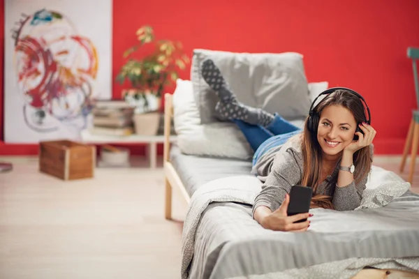 Schöne lächelnde Brünette auf dem Bauch auf dem Bett im Schlafzimmer liegend, Musik hörend und Smartphone benutzend. — Stockfoto