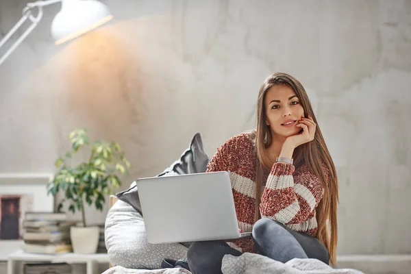 Beautiful smiling Caucasian brunette sitting on the bed in bedroom, looking at camera and using laptop. — Stock Photo, Image