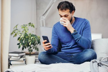 Çekici gülümseyen Arap adam rahat yatak odası, yatakta oturup kahve içme ve akıllı telefon mesaj attığın için kullanarak giyinmiş.