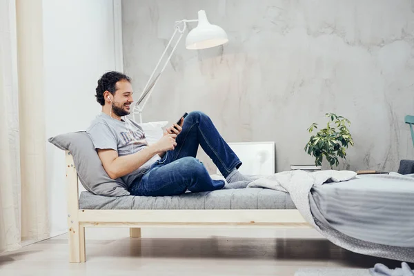 Hombre árabe sonriente sentado en la cama en el dormitorio y demandando teléfono inteligente. En los oídos auriculares . —  Fotos de Stock