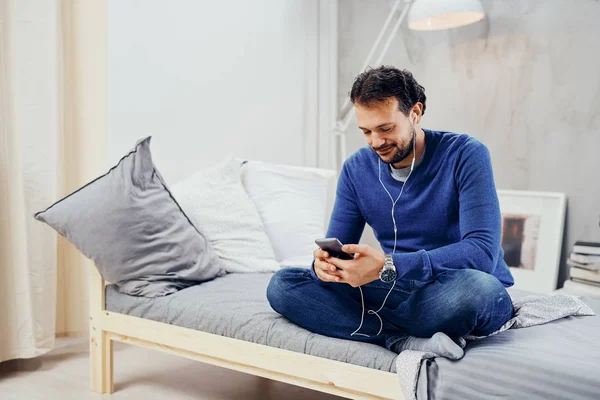 Felice uomo arabo carino seduto sul letto in camera da letto con le gambe incrociate e ascoltare musica su smart phone . — Foto Stock