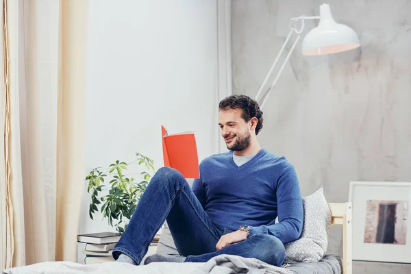 Vrolijke schattig Arabische man gekleed casual boek lezen en ontspannen zittend op het bed in de slaapkamer. — Stockfoto