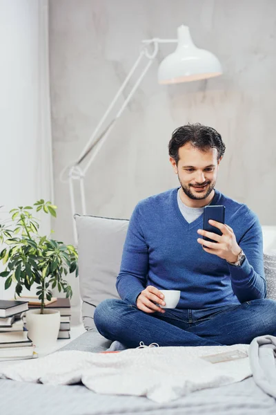 Atractivo hombre árabe sonriente vestido casual sentado en la cama en el dormitorio, beber café y usar el teléfono inteligente para enviar mensajes de texto . —  Fotos de Stock