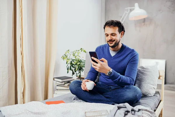 Aantrekkelijke lachende Arabische man gekleed casual zittend op bed in de slaapkamer, drinken koffie en met behulp van slimme telefoon voor het texting. — Stockfoto