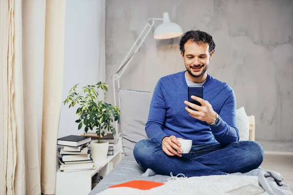 Aantrekkelijke lachende Arabische man gekleed casual zittend op bed in de slaapkamer, drinken koffie en met behulp van slimme telefoon voor het texting. — Stockfoto
