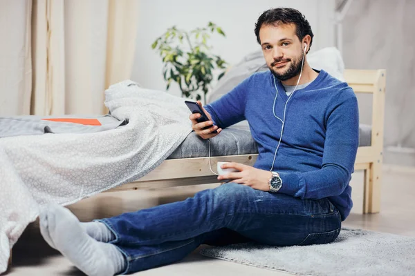 Hombre árabe atractivo sentado en el suelo en el dormitorio, escuchando música a través del teléfono inteligente y tomando café mientras mira la cámara . —  Fotos de Stock