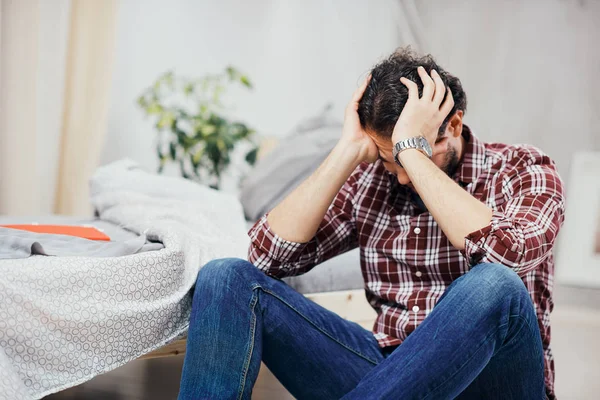 Attractive arab man in jeans and plaid shirt sitting on the floor in bedroom and holding head. Men's issues concept. — Stock Photo, Image