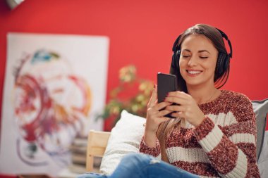 Kulaklara kulaklıklarla yatağa oturmuş ve akıllı telefon kullanan genç beyaz esmer. Mercan kırmızı arka plan.