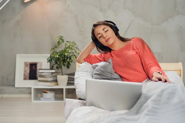 Hermosa morena caucásica escuchando música y usando el portátil mientras está sentada en la cama en el dormitorio . — Foto de Stock