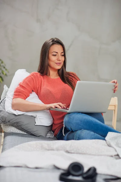 Gyönyörű kaukázusi barna ül az ágyon a hálószobában, és használ laptop. — Stock Fotó