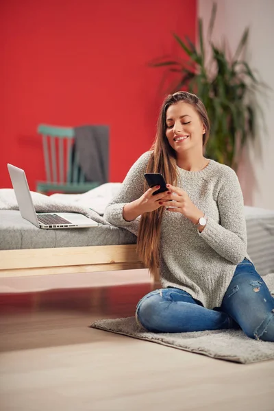 Hermosa morena caucásica vestida casual sentada en el suelo y apoyada en la cama en el dormitorio mientras habla por teléfono. En la cama portátil . — Foto de Stock