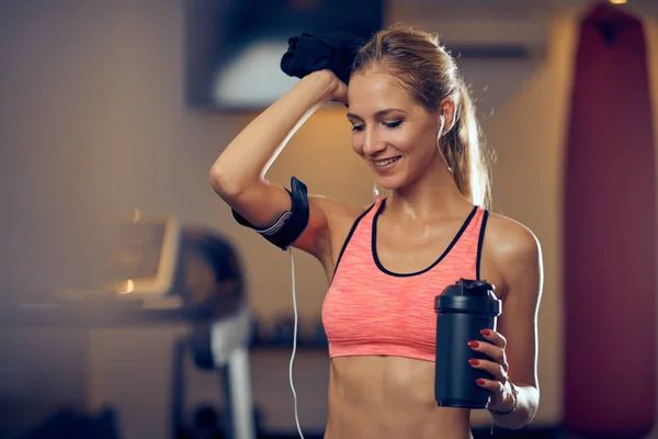 Joven mujer atlética sonriente caucásica en ropa deportiva sosteniendo botella de agua y limpiando el sudor mientras está de pie en el gimnasio . — Foto de Stock