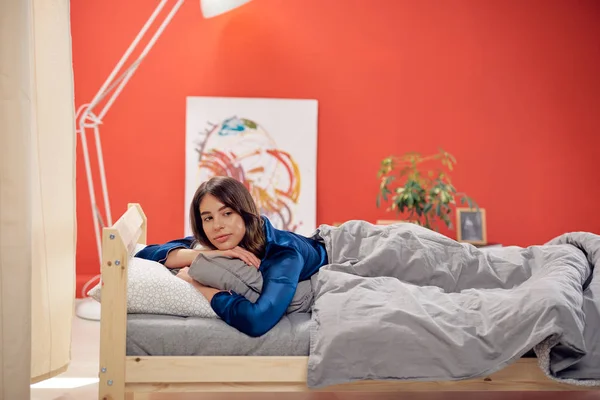 Atractiva morena caucásica vestida con pijamas azules acostada en la cama sobre el estómago. Hora de la mañana . — Foto de Stock