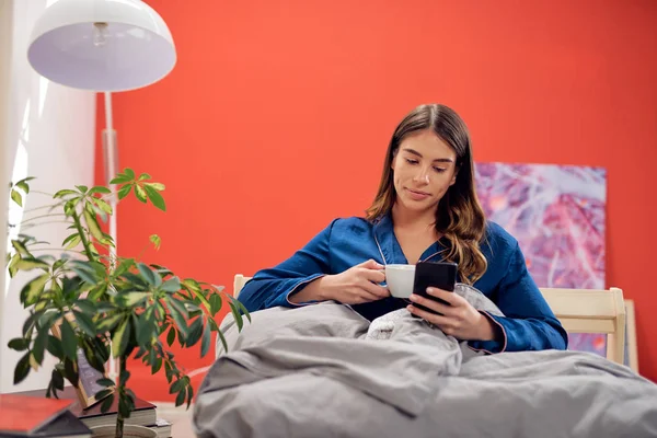 Bonita morena caucásica vestida con pijamas azules sentada en la cama por la mañana, bebiendo café y usando un teléfono inteligente . — Foto de Stock