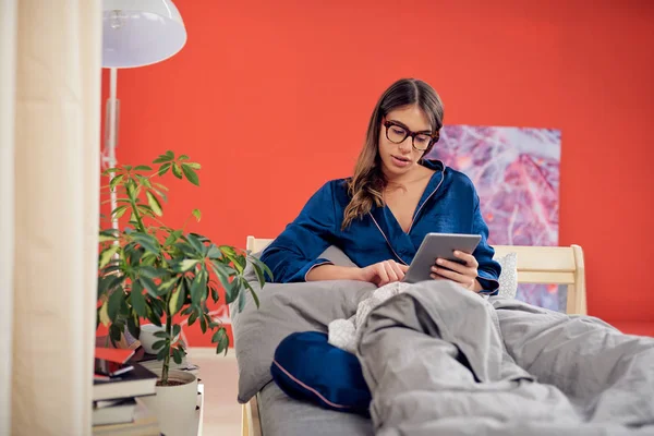 Πανέμορφο Καυκάσιος μελαχρινή με γυαλιά και ντυμένος με μπλε πιτζάμες κάθεται στο κρεβάτι στο υπνοδωμάτιο, το πρωί και χρησιμοποιώντας tablet. — Φωτογραφία Αρχείου