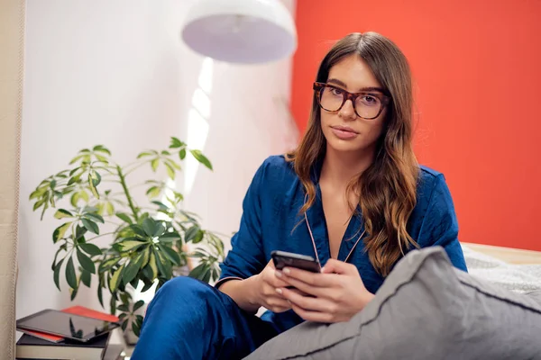 Affascinante bruna caucasica vestita in pigiama blu e con gli occhiali che guardano la macchina fotografica mentre si siede sul letto in bedrooma e usando lo smartphone. Ora del mattino . — Foto Stock