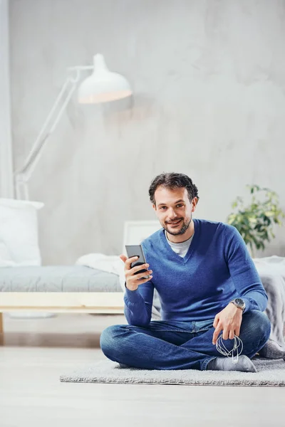 Aantrekkelijke Arabische man gekleed casual zittend op de vloer in de slaapkamer en met behulp van slimme telefoon voor lezen of verzenden van bericht. — Stockfoto