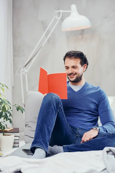 Cheerful cute arab man dressed casual reading book and relaxing while sitting on the bed in bedroom. — Stock Photo, Image