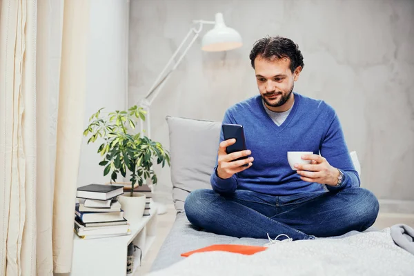 Привлекательный улыбающийся араб одет непринужденно, сидит на кровати в спальне, пьет кофе и использует смартфон для смс . — стоковое фото