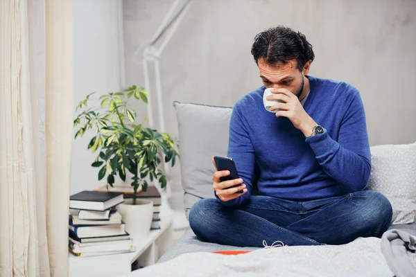 Attractive arab man dressed casual sitting on bed in bedroom, drinking coffee and using smart phone for texting. — Stock Photo, Image