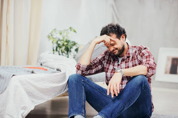 Attractive arab man in jeans and plaid shirt sitting on the floor in bedroom and holding head. Men's issues concept. — Stock Photo, Image