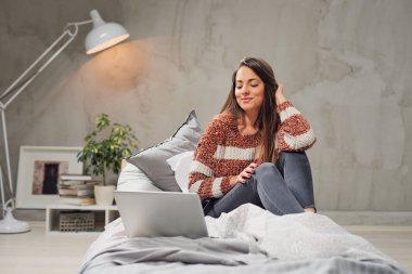 Güzel beyaz esmer yatak odasında yatağın üstüne oturan ve dizüstü bilgisayar kullanarak.