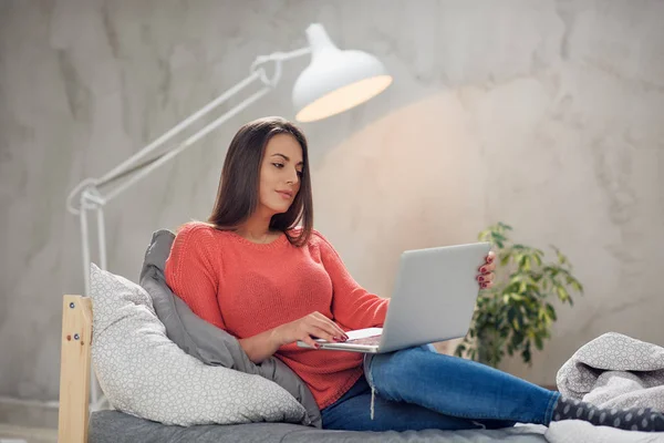 Schöne kaukasische Brünette sitzt auf dem Bett im Schlafzimmer und mit Laptop. — Stockfoto