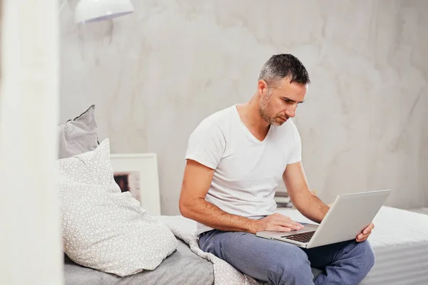 Szép kaukázusi középső férfi öltözött alkalmi használ laptop, miközben ül ágyas hálószoba. — Stock Fotó