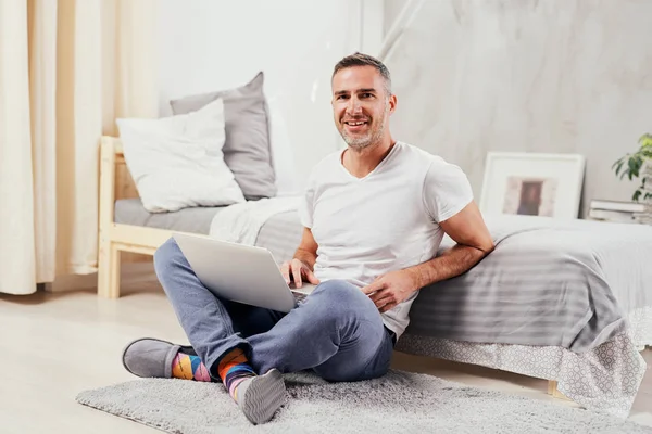 Handsome middle aged man sitting on the floor wirh crossed legs, leaning on bed and using laptop. — Stock Photo, Image