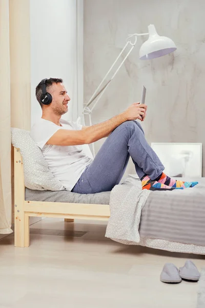 Sorridente uomo caucasico di mezza età vestito casual rilassante sul letto in camera da letto, ascoltando musica e utilizzando tablet . — Foto Stock