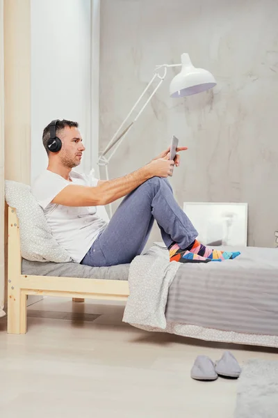 Кавказский мужчина среднего возраста одет непринужденно расслабляясь на кровати в спальне, слушая музыку и используя планшет . — стоковое фото