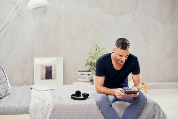 Kaukasische man in veertig zittend op bed in de slaapkamer en het gebruik van Tablet PC. — Stockfoto