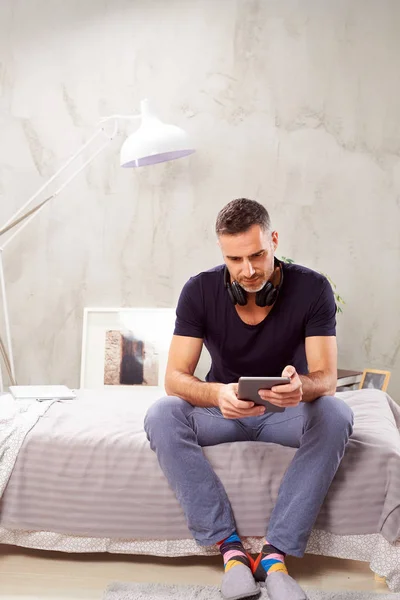 Кавказский мужчина в сорок лет сидит на кровати в спальне и с помощью планшета . — стоковое фото