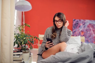 Seksi Caucasain gözlük ile esmer ve sabah yatakta oturuyor ve akıllı telefon kullanarak Kazak giymiş.