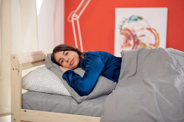 Привлекательная брюнетка-кавказка в голубой пижаме лежит в постели на животе. Утреннее время . — стоковое фото