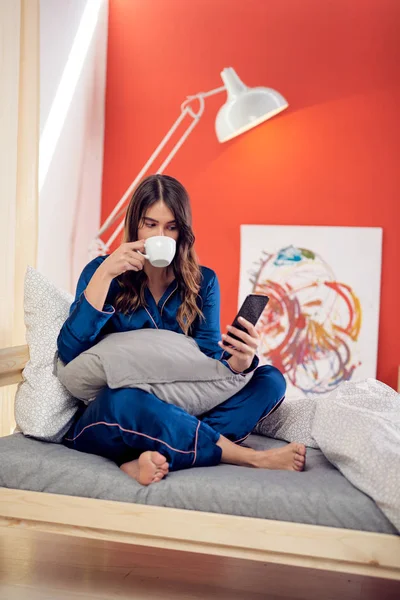 Gorgeous brunetka w piżamie niebieski, siedząc na łóżku w sypialni z skrzyżowanymi nogami, picie porannej kawy i przy użyciu smartfona. — Zdjęcie stockowe