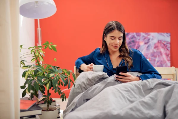 Bella bruna caucasica vestita in pigiama blu seduta a letto al mattino, che beve caffè e utilizza lo smartphone . — Foto Stock