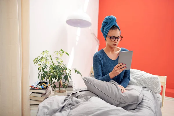 Hermosa morena con anteojos y toalla en la cabeza usando tableta mientras está sentada en la cama en el dormitorio . — Foto de Stock