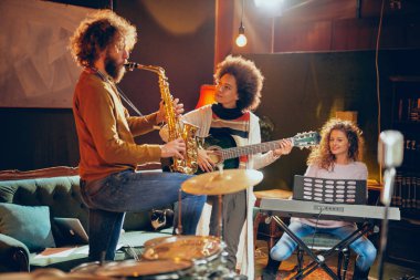 Karışık ırk kadın adam saksafon oynamaya akustik gitar çalmak. Ev stüdyosu iç. Clavier oynarken arka plan kadında.