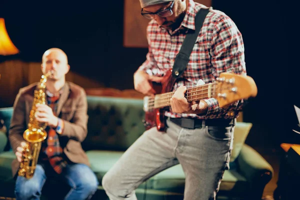 Young Caucasian man with hat on head playing bass guitar while standing in home studio. In background saxophonist playing his instrument. — Stock Photo, Image