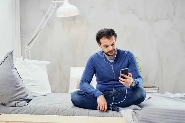 Happy cute arab man sitting on the bed in bedroom with legs crossed and listening music over smart phone. — Stock Photo, Image