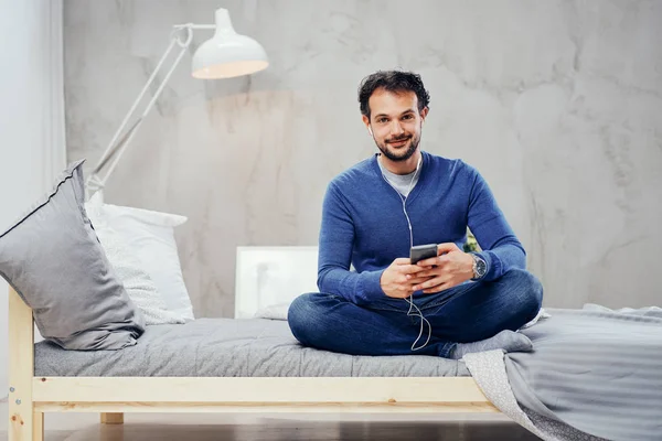 Felice uomo arabo carino seduto sul letto in camera da letto con le gambe incrociate e ascoltare musica su smart phone . — Foto Stock
