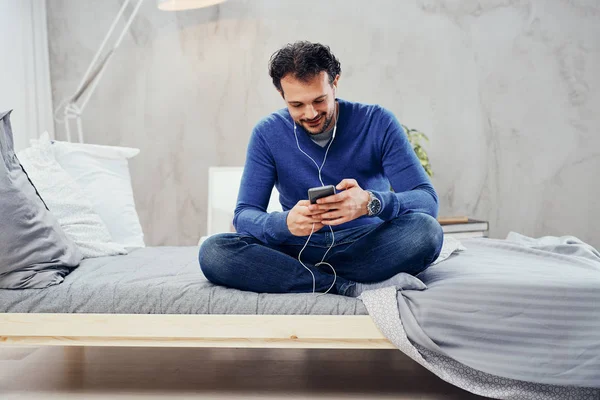 Feliz hombre árabe lindo sentado en la cama en el dormitorio con las piernas cruzadas y escuchar música a través del teléfono inteligente . —  Fotos de Stock