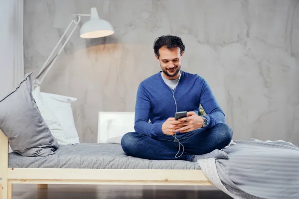 Gelukkig schattig Arabische man zittend op het bed in de slaapkamer met de benen gekruist en luisteren van muziek via smartphone. — Stockfoto