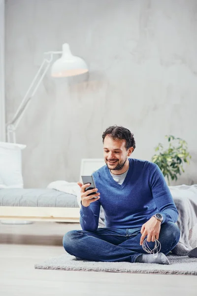 Aantrekkelijke Arabische man gekleed casual zittend op de vloer in de slaapkamer en met behulp van slimme telefoon voor lezen of verzenden van bericht. — Stockfoto