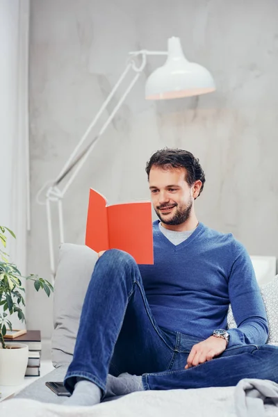 Cheerful cute arab man dressed casual reading book and relaxing while sitting on the bed in bedroom. — Stock Photo, Image