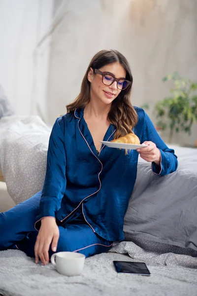 Junge kaukasische Brünette im Pyjama trinkt Kaffee, hält Teller mit Croissant und sitzt morgens auf dem Boden im Schlafzimmer. — Stockfoto