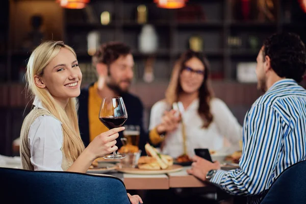 Retrato de la hermosa mujer rubia caucásica sentada en el restaurante y sosteniendo una copa de vino tinto. En segundo plano amigos cenando . —  Fotos de Stock