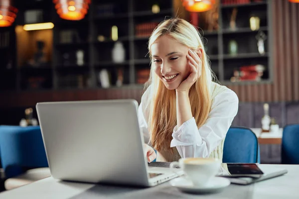 Linda sorridente branco loira blogueira feminina vestida inteligente casual sentado na cafetaria e usando laptop . — Fotografia de Stock